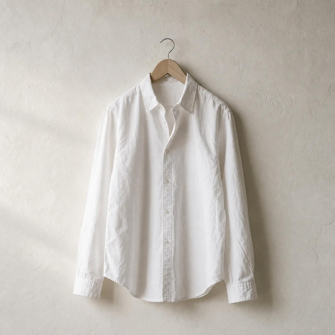 A white cotton shirt on a plain wooden hanger against a bare plaster wall, raking light across the cloth